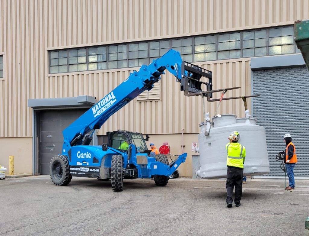 Genie telehandler lifting HVAC chamber section in front of a building with workers